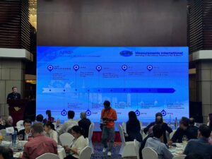 Measurements International presentation at an electrical metrology conference, with attendees seated before a large display screen.